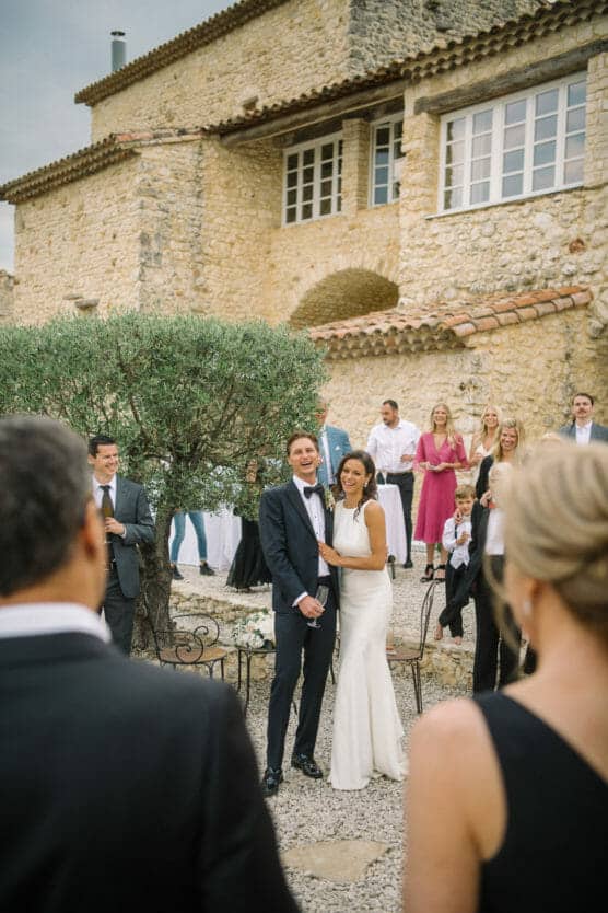A bride and groom smile and laugh together outdoors in front of a stone building, surrounded by elegantly dressed guests at a wedding celebration. by Justin Salem Meyer
