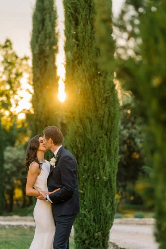 A bride in a white dress and a groom in a dark suit kiss in a garden at sunset, surrounded by tall green trees and soft golden light. by Justin Salem Meyer