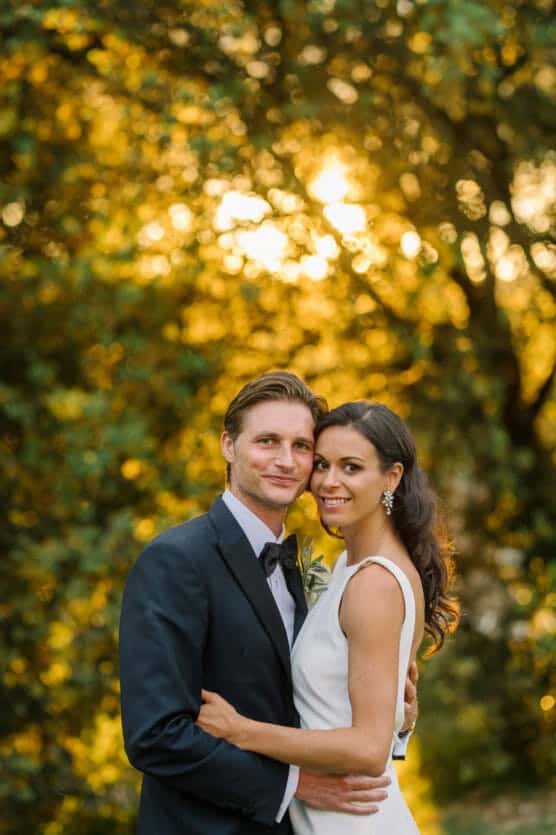 A couple dressed in formal wedding attire embrace and smile at the camera outdoors, with golden sunlight and green foliage in the background. by Justin Salem Meyer