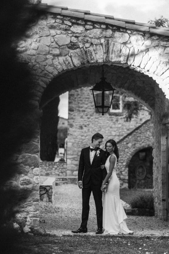 A bride and groom stand close together under a stone archway outside a rustic building, holding hands and gazing at each other. The bride wears a long white dress; the groom is in a black tuxedo. The photo is black and white. by Justin Salem Meyer