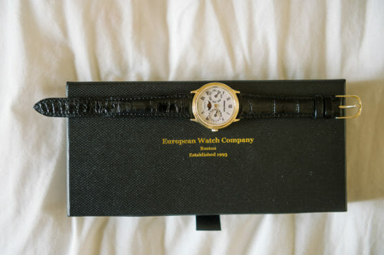A gold wristwatch with a black leather strap is displayed on top of a black textured box labeled European Watch Company Boston Established 1993. The background is a white fabric surface. by Justin Salem Meyer