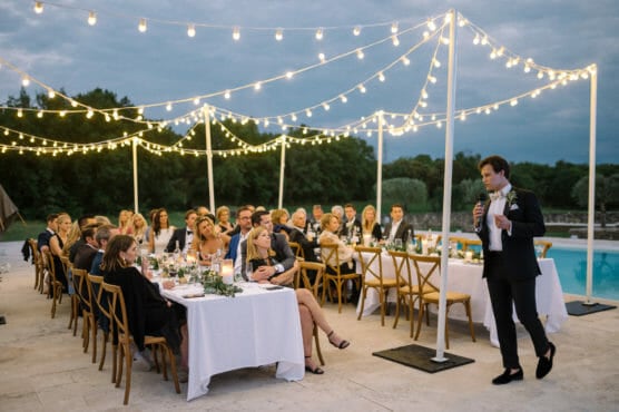 A man in a suit speaks into a microphone beside a pool at an outdoor evening event. Guests sit at tables decorated with white cloths and string lights hang overhead, creating a festive atmosphere. by Justin Salem Meyer