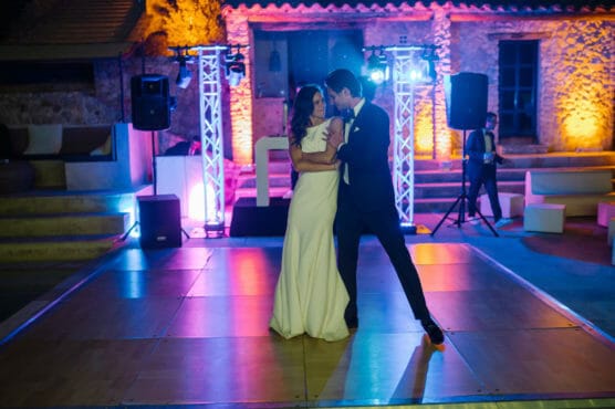A bride and groom dance closely on a lit-up dance floor at night, surrounded by colorful lights and stone buildings, creating a festive and romantic atmosphere. by Justin Salem Meyer