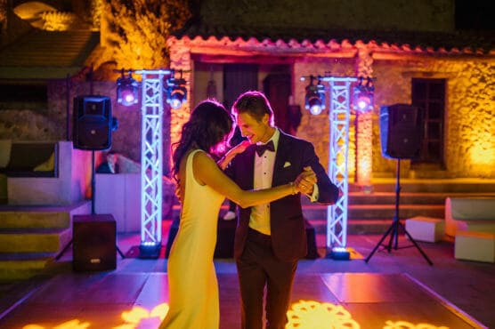 A couple dressed formally, with the woman in a white dress and the man in a suit, dance together on an outdoor dance floor at night, illuminated by colorful stage lights and surrounded by stone buildings. by Justin Salem Meyer