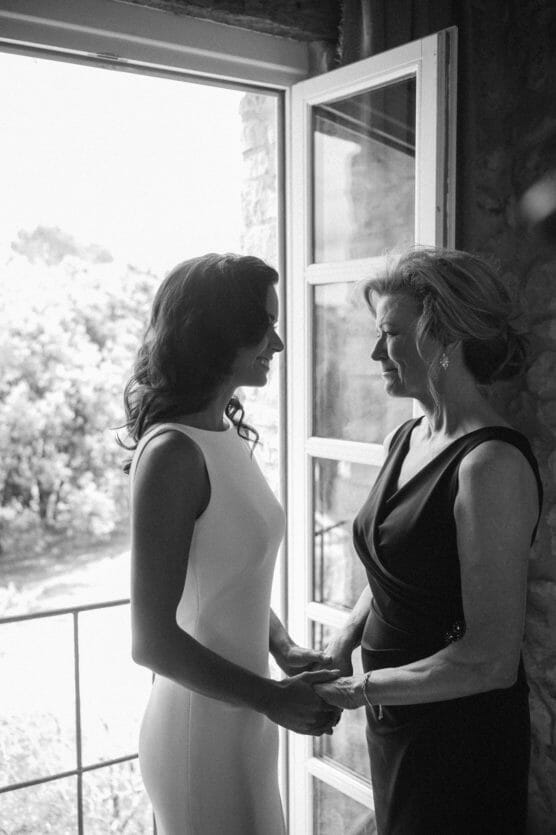 A bride in a white dress and a woman in a dark dress stand facing each other, holding hands and smiling, by a window with sunlight streaming in, in a softly lit room. by Justin Salem Meyer