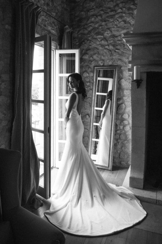 A woman in a sleeveless, floor-length wedding dress stands by an open door, looking back and smiling. Her reflection is visible in a tall mirror beside her in a rustic room with stone walls. by Justin Salem Meyer