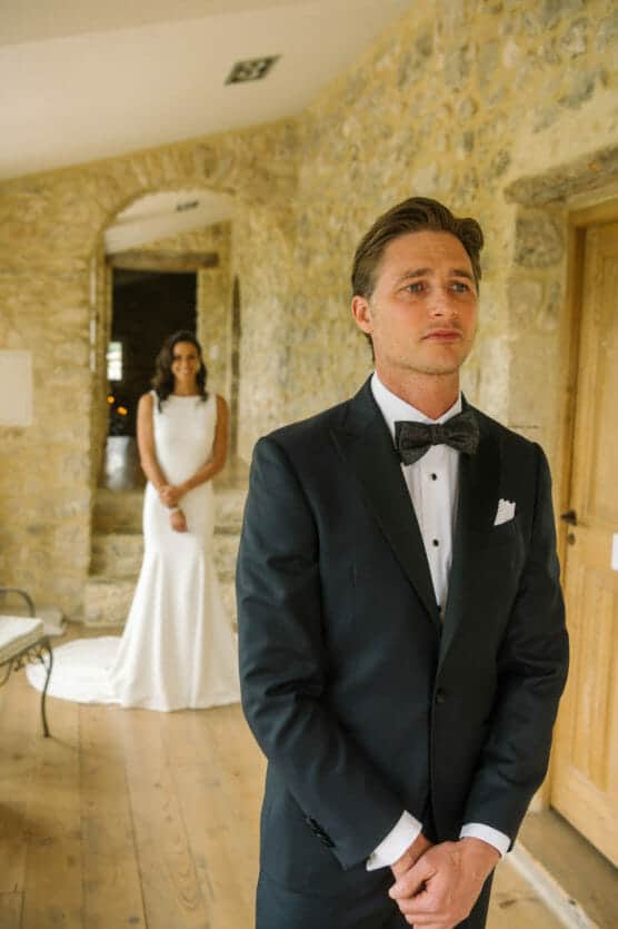 A groom in a black tuxedo stands in the foreground, looking emotional, while a bride in a white dress stands behind him, both indoors against a stone wall background. by Justin Salem Meyer
