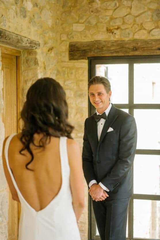 A man in a black tuxedo smiles warmly at a woman in a white, backless dress. They stand indoors near a stone wall and large window, suggesting a special or formal occasion. by Justin Salem Meyer