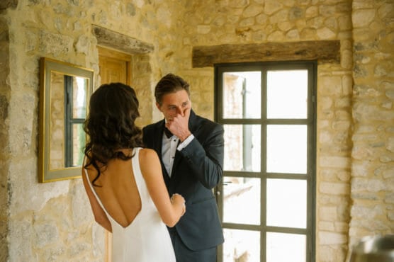 A woman in a white dress holds hands with a man in a suit, who appears emotional and is wiping his eyes, inside a stone-walled room with large windows and a mirror on the wall. by Justin Salem Meyer
