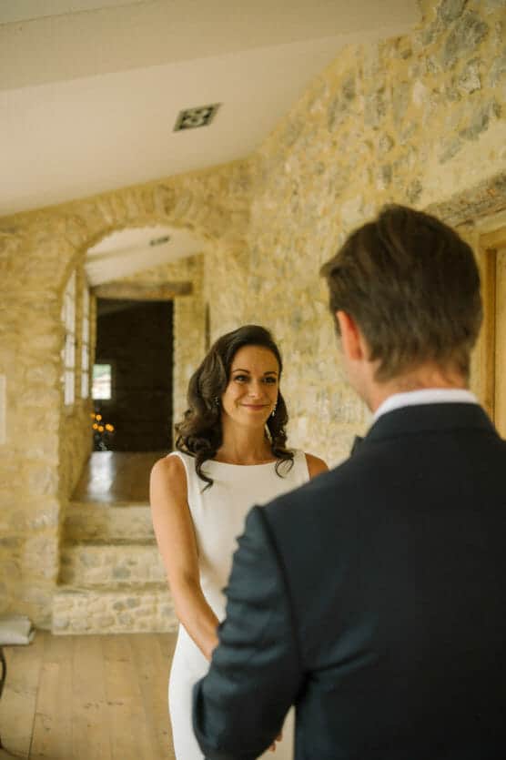 A bride in a white dress smiles warmly at a groom in a dark suit. They stand indoors by a stone wall, holding hands, sharing an intimate moment together. by Justin Salem Meyer