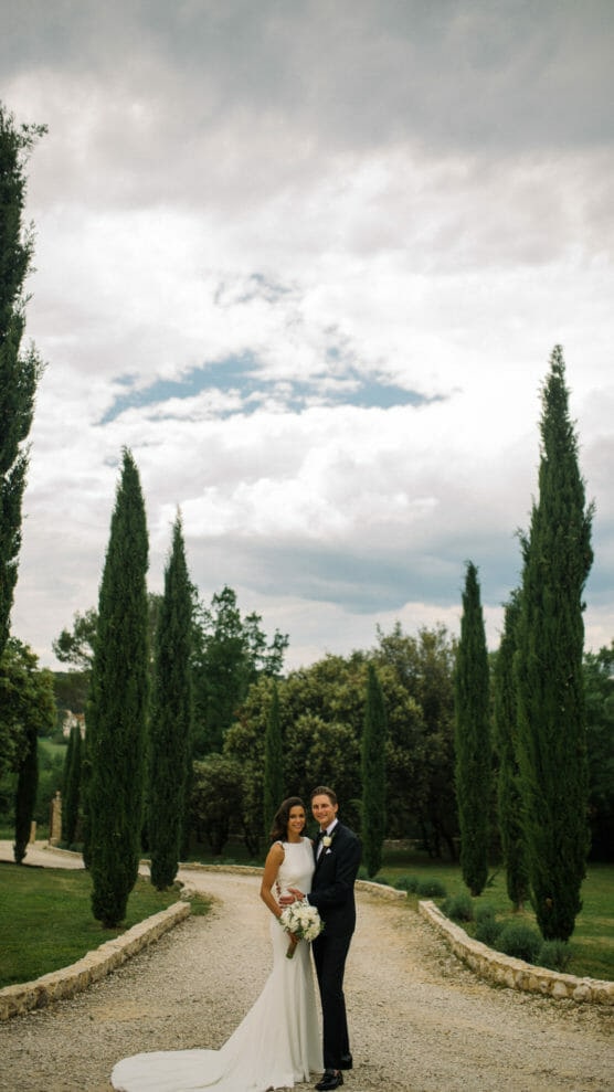A bride and groom stand together on a gravel path lined with tall cypress trees, surrounded by lush greenery under a cloudy sky. The bride wears a white dress and holds a bouquet; the groom wears a dark suit. by Justin Salem Meyer