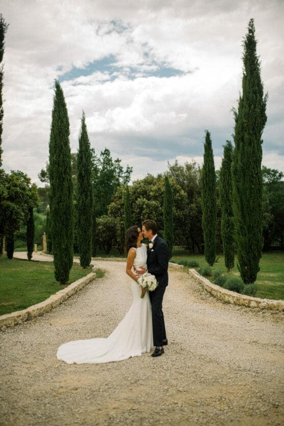 A bride and groom share a kiss on a gravel path lined with tall cypress trees and greenery, under a partly cloudy sky. The bride wears a long white dress and holds a bouquet. by Justin Salem Meyer