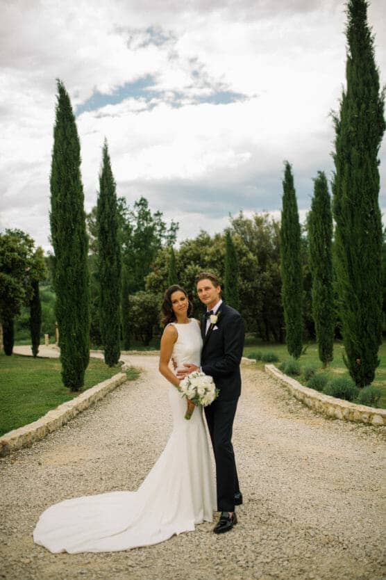 A bride in a long white dress and a groom in a black tuxedo stand together on a gravel path lined with tall cypress trees, with greenery and a cloudy sky in the background. by Justin Salem Meyer