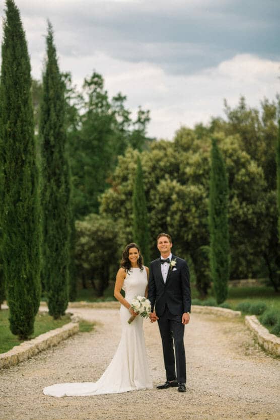 A bride in a white dress and a groom in a black tuxedo stand together on a gravel path lined with tall green trees, holding hands and smiling outdoors. by Justin Salem Meyer