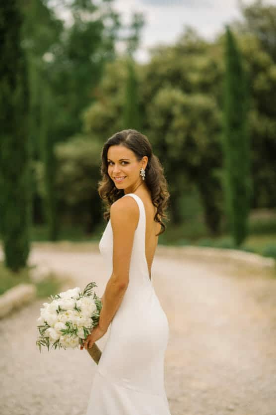 A woman in a white sleeveless wedding dress stands on a gravel path, holding a bouquet of white flowers and smiling over her shoulder, with trees and greenery in the background. by Justin Salem Meyer