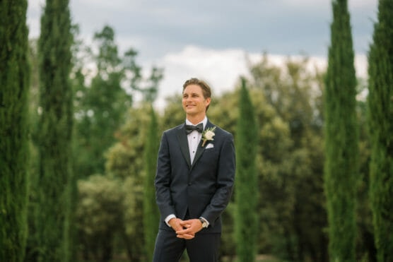 A man in a dark suit with a bow tie and boutonnière stands outdoors among tall green trees, smiling and clasping his hands, with a soft, natural background. by Justin Salem Meyer