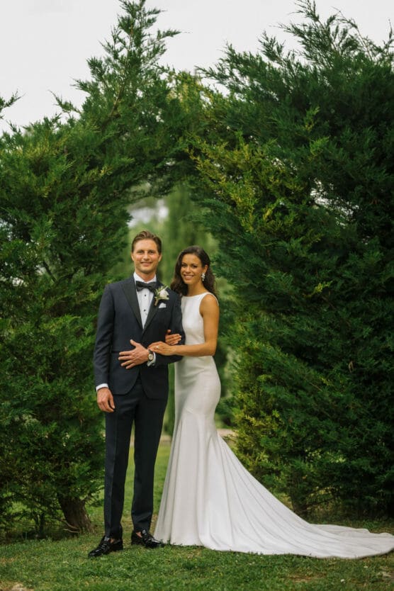 A bride in a fitted white gown and a groom in a black tuxedo stand together, smiling, beneath a natural arch formed by lush green trees outdoors. by Justin Salem Meyer