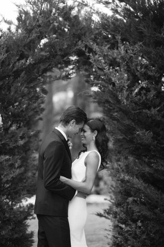 A couple stands under an archway of tall greenery, facing each other closely and smiling. The man wears a suit and the woman wears a sleeveless dress, suggesting a wedding or special occasion. The image is in black and white. by Justin Salem Meyer