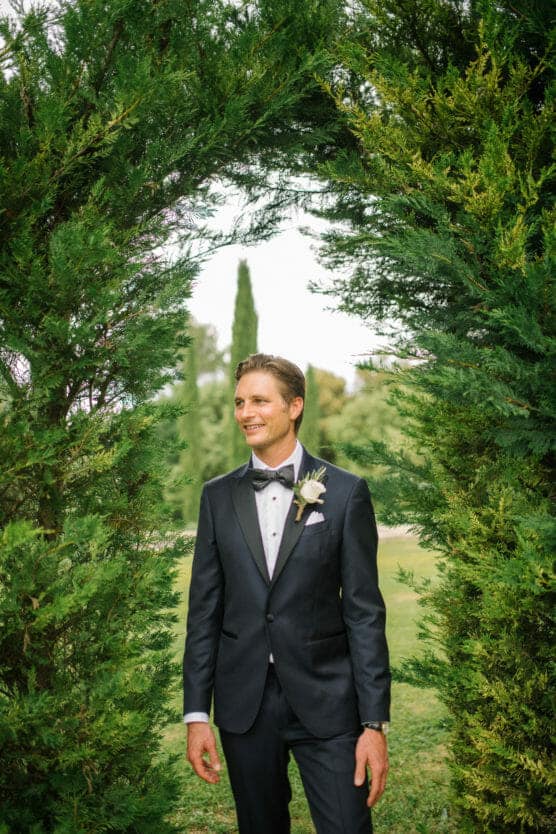 A man in a dark suit and bow tie stands smiling under a green arch of tall, leafy bushes in an outdoor setting, with grass and trees visible in the background. by Justin Salem Meyer