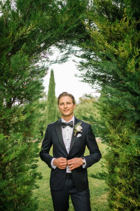 A groom in a black tuxedo with a bow tie stands outdoors, framed by green trees or bushes, smiling confidently with hands adjusting his jacket. by Justin Salem Meyer