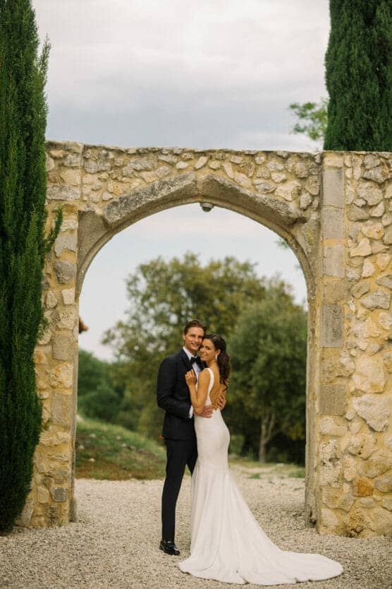 A bride in a white gown and a groom in a dark suit embrace and smile under a stone arch outdoors, surrounded by greenery and trees. by Justin Salem Meyer