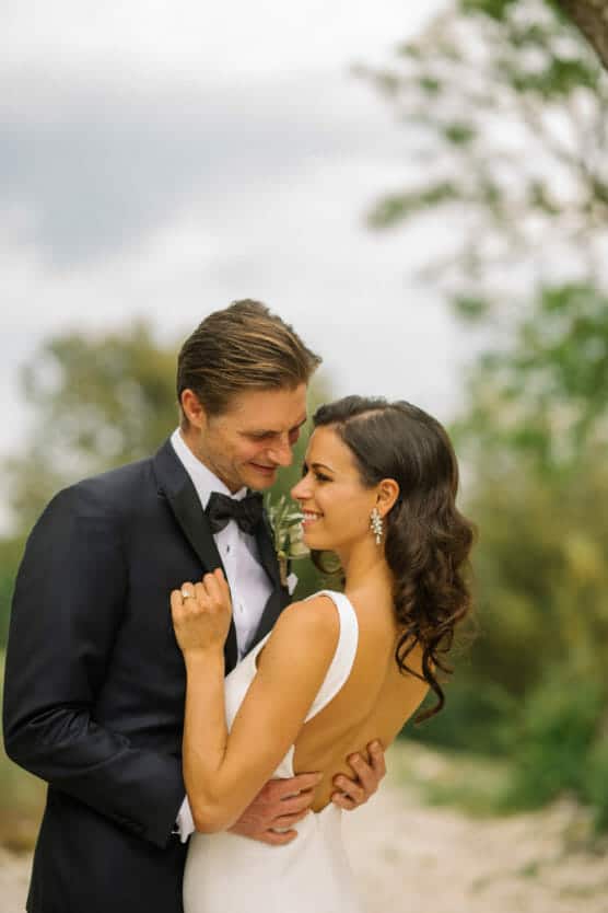 A bride in a white dress and a groom in a black tuxedo embrace outdoors, smiling at each other lovingly with greenery and a cloudy sky in the background. by Justin Salem Meyer