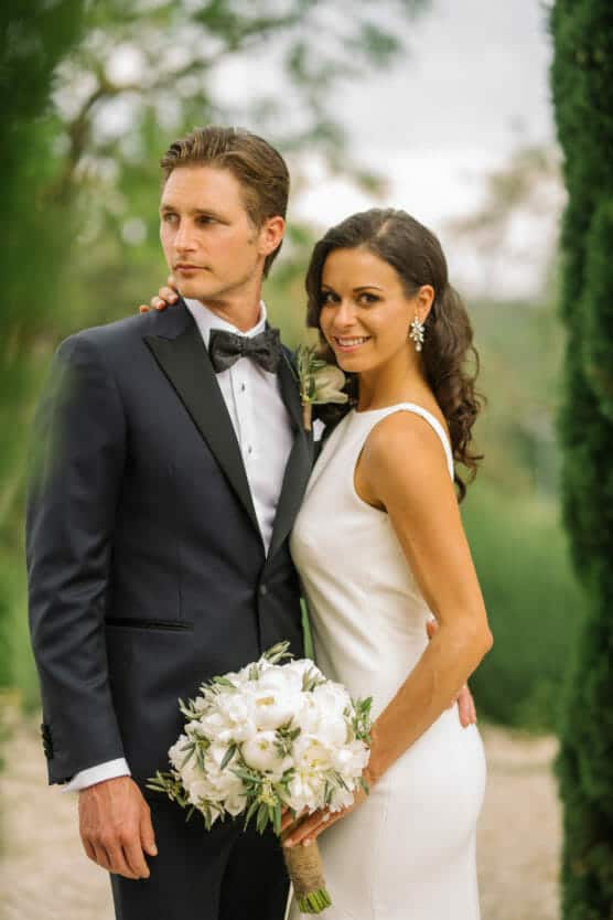 A bride in a white dress holds a white bouquet and smiles while standing next to a groom in a black tuxedo outdoors, surrounded by greenery. by Justin Salem Meyer