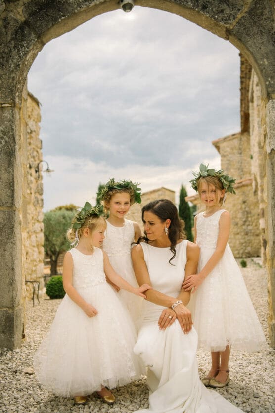 A bride in a white dress kneels and smiles, surrounded by three young flower girls in matching white dresses and leaf crowns, outdoors under a stone archway. by Justin Salem Meyer