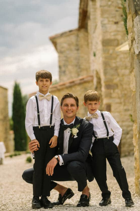 A man in a tuxedo kneels outdoors between two young boys wearing white shirts, black pants, suspenders, and bow ties. They all smile at the camera in front of stone buildings on a sunny day. by Justin Salem Meyer