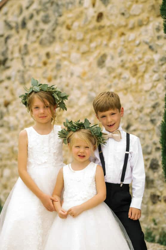 Three children dressed in formal attire stand in front of a stone wall. Two girls wear white dresses and leafy crowns; a boy wears a white shirt with a bow tie and black suspenders. All are smiling. by Justin Salem Meyer