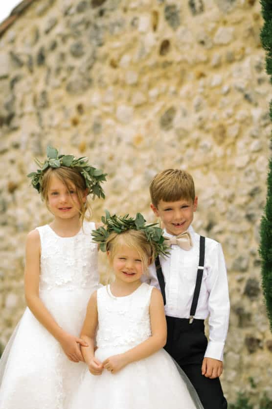 Three young children dressed for a wedding pose in front of a stone wall. Two girls wear white dresses and leafy headbands, and a boy wears a white shirt with a bow tie and black suspenders. All are smiling. by Justin Salem Meyer