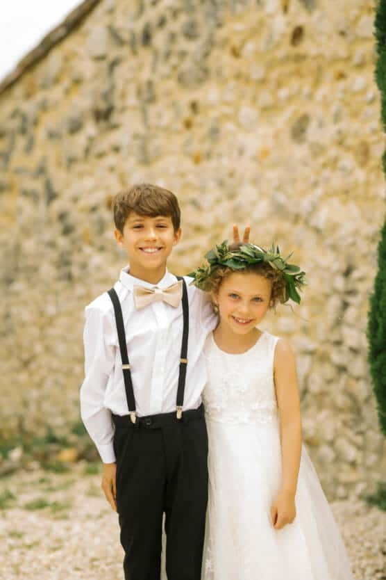 A young boy in suspenders and a bow tie stands smiling next to a girl in a white dress and floral crown. The boy playfully gives her bunny ears. They are outdoors, in front of a stone wall. by Justin Salem Meyer