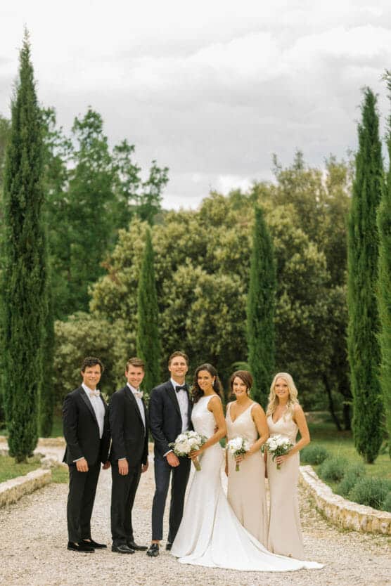 Five people stand outdoors on a gravel path lined with tall trees. The two men and three women are dressed formally, with the women holding bouquets, suggesting a wedding party. Lush greenery and cloudy skies are in the background. by Justin Salem Meyer