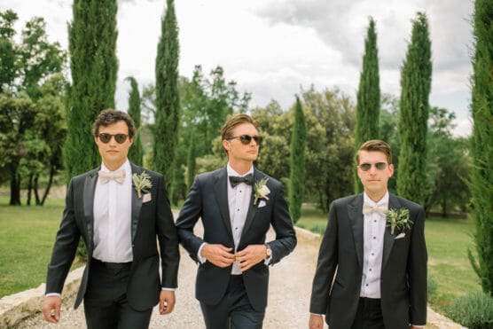 Three men in tuxedos and sunglasses walk confidently down a path lined with trees. They have boutonnières on their jackets and a natural, green outdoor setting surrounds them. The sky is partly cloudy. by Justin Salem Meyer