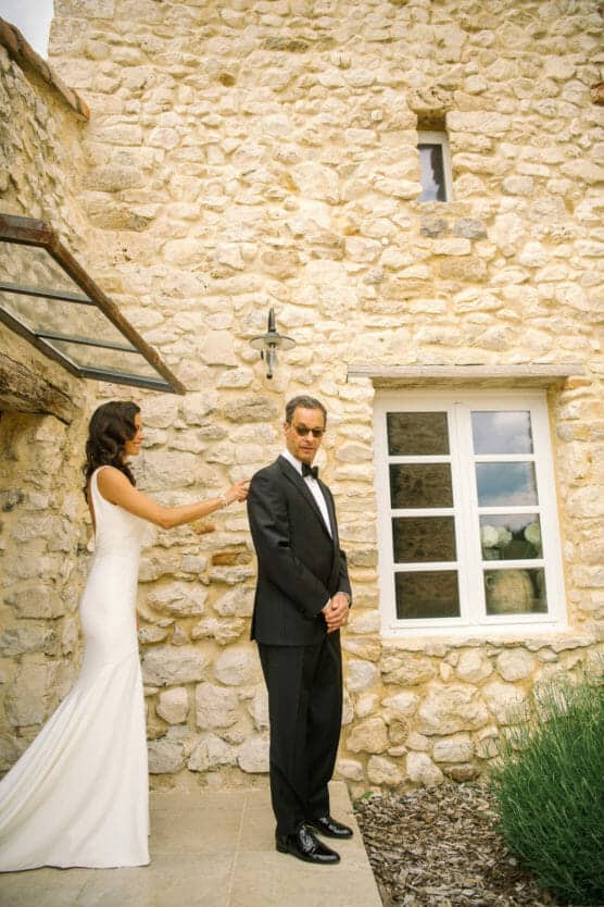 A bride in a white dress approaches a man in a tuxedo, touching his shoulder from behind outside a rustic stone building with white-framed windows. The man stands facing away, appearing unaware of her presence. by Justin Salem Meyer