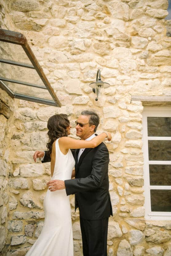 A bride in a white dress hugs an older man in a black suit and sunglasses, both smiling, standing outside against a rustic stone wall with a window and light fixture. by Justin Salem Meyer