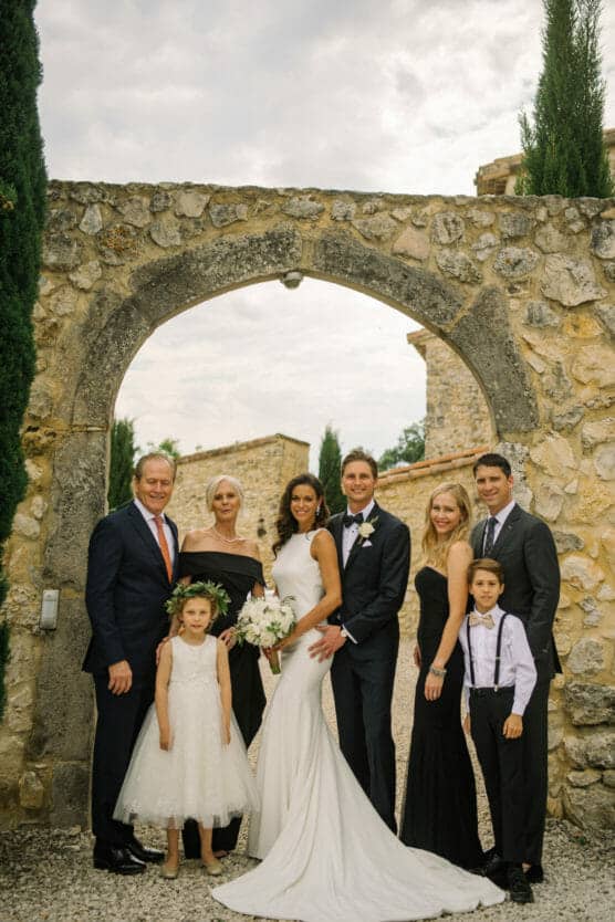A bride and groom stand under a stone archway, surrounded by four adults and two children dressed formally, posing together outdoors on a cloudy day. by Justin Salem Meyer