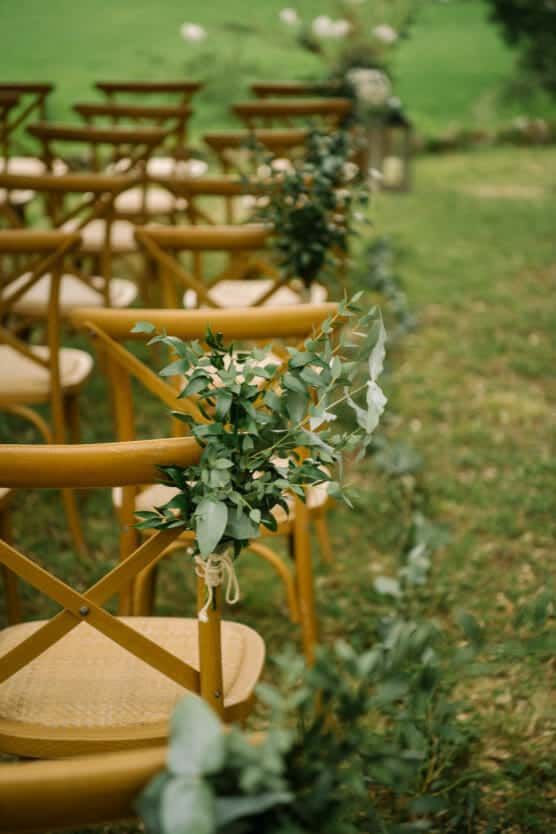 Rows of wooden chairs with cross backs are arranged outdoors on grass, decorated with green leafy foliage, creating a natural and elegant setting, likely for a wedding ceremony. by Justin Salem Meyer