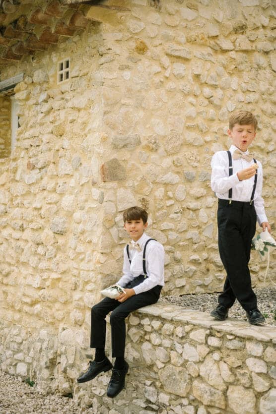 Two young boys in white shirts, black pants, and suspenders eat snacks in front of a rustic stone wall. One boy sits on a ledge while the other stands beside him, both appearing relaxed and playful. by Justin Salem Meyer