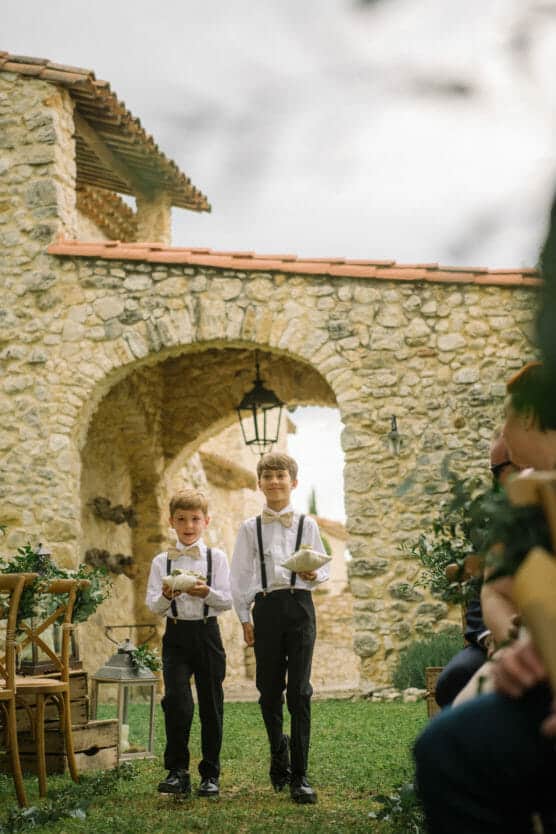 Two young boys dressed in white shirts, black pants, and suspenders walk down an outdoor aisle at a rustic stone venue, each holding a pillow. Guests are seated on either side, and greenery decorates the chairs. by Justin Salem Meyer