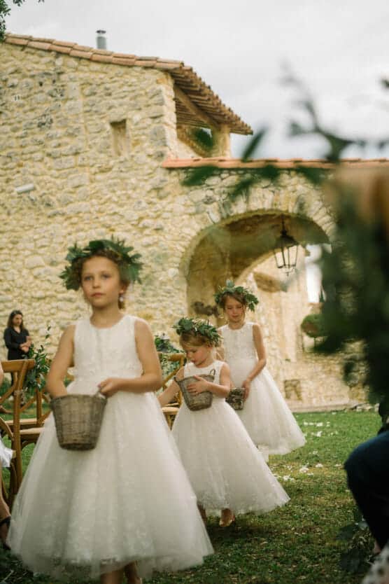 Three young flower girls in white dresses and green flower crowns walk with baskets down an outdoor aisle beside a stone building at a wedding ceremony. by Justin Salem Meyer