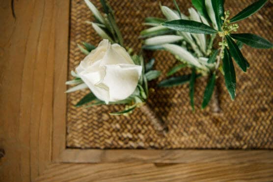 A white rose with green leaves rests on a woven placemat beside another small floral arrangement, all set on a wooden table. by Justin Salem Meyer