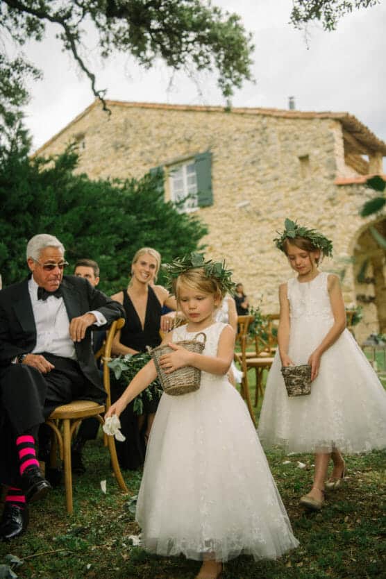 Two young flower girls in white dresses and floral crowns walk down an outdoor aisle, scattering petals from baskets. Seated wedding guests watch, including an older man in a tuxedo with pink-striped socks. A stone house is in the background. by Justin Salem Meyer