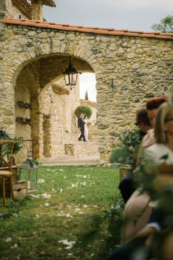A bride and groom walk hand in hand through an arched stone passageway at an outdoor wedding, with guests seated on either side and flower petals scattered on the grass. by Justin Salem Meyer