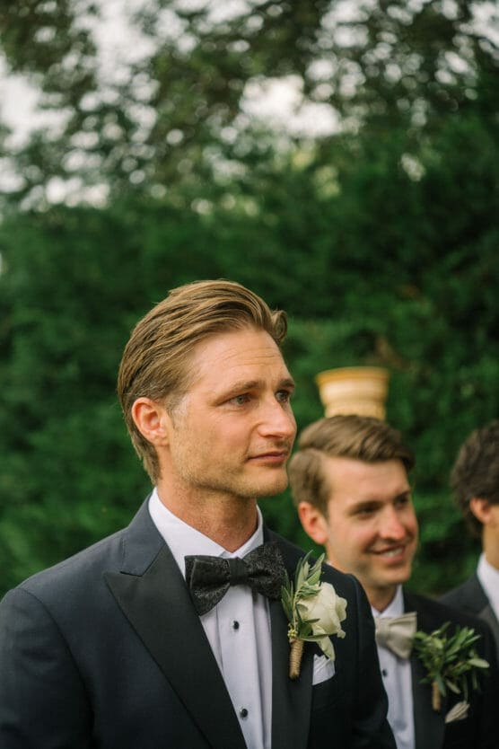 A man in a black tuxedo with a black bow tie and white boutonnière stands outdoors, looking ahead. Two other men in tuxedos are behind him, all surrounded by greenery. by Justin Salem Meyer