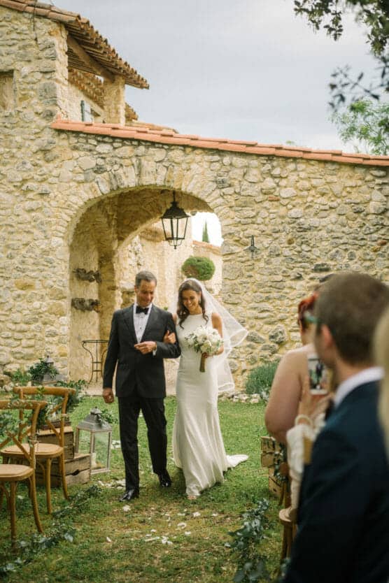 A bride in a white dress walks arm in arm with an older man in a suit down an outdoor aisle lined with chairs and greenery, beside a stone building with arches. Guests watch the procession. by Justin Salem Meyer