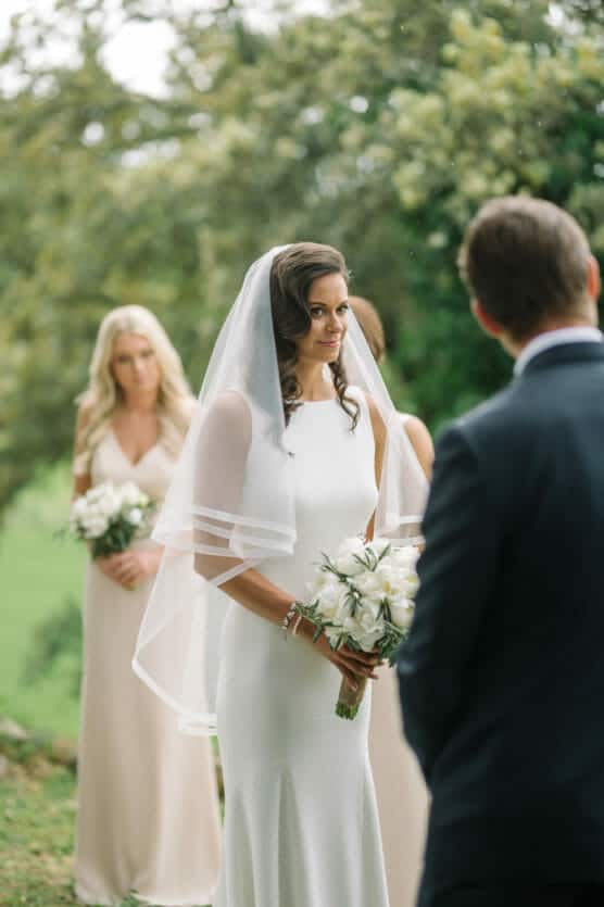 A bride in a white dress and veil holding a bouquet stands outdoors, looking at a man in a suit. Bridesmaids in light dresses holding flowers are in the background, with greenery all around. by Justin Salem Meyer