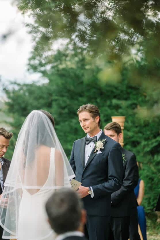 A groom in a black tuxedo reads vows to a bride in a white dress and veil during an outdoor wedding ceremony, surrounded by greenery and wedding guests. by Justin Salem Meyer
