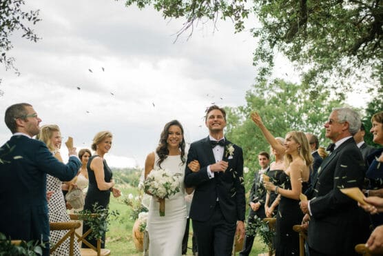 A bride in a white dress and groom in a tuxedo walk arm in arm down an outdoor aisle, smiling, surrounded by cheering guests who are tossing flower petals. Trees and greenery are visible in the background. by Justin Salem Meyer
