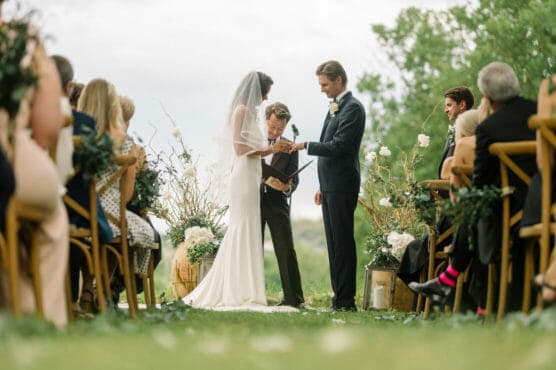 A bride and groom stand together outdoors during their wedding ceremony, holding hands as an officiant reads from a book. Guests are seated on either side, watching the couple. by Justin Salem Meyer
